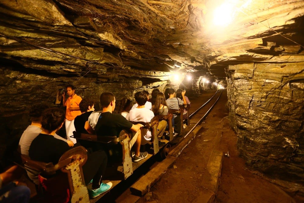 Grupo de pessoas em passeio de carrinho por túnel iluminado dentro de mina de ouro em Ouro Preto, com guia explicando a história do local